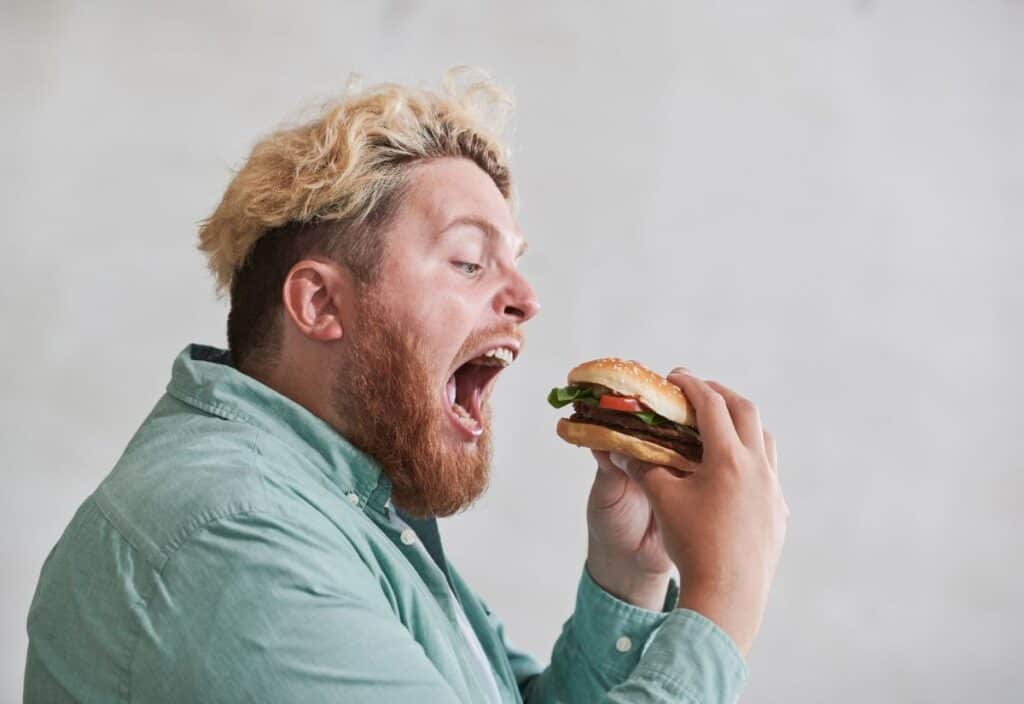 A man with blonde hair and a beard is wearing a green shirt and holding a burger with an open mouth, about to take a bite.