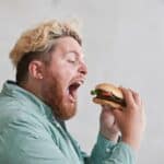 A man with blonde hair and a beard is wearing a green shirt and holding a burger with an open mouth, about to take a bite.