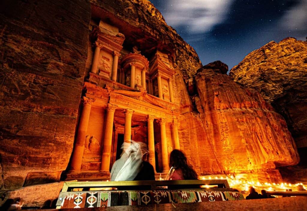 People in front of the illuminated facade of Al-Khazneh in Petra at night, with a starry sky overhead.