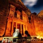 People in front of the illuminated facade of Al-Khazneh in Petra at night, with a starry sky overhead.