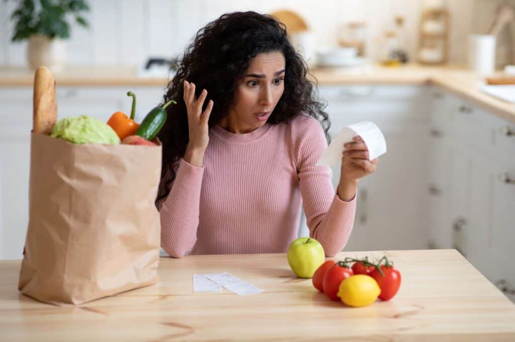 Woman in a kitchen looks surprised at a receipt. A paper bag with groceries, including bread and vegetables, is on the table.