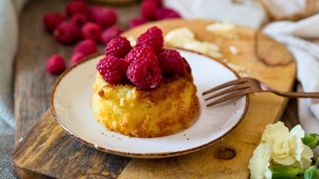 A small cake topped with fresh raspberries sits on a plate with a fork. More raspberries are scattered on the wooden surface nearby.