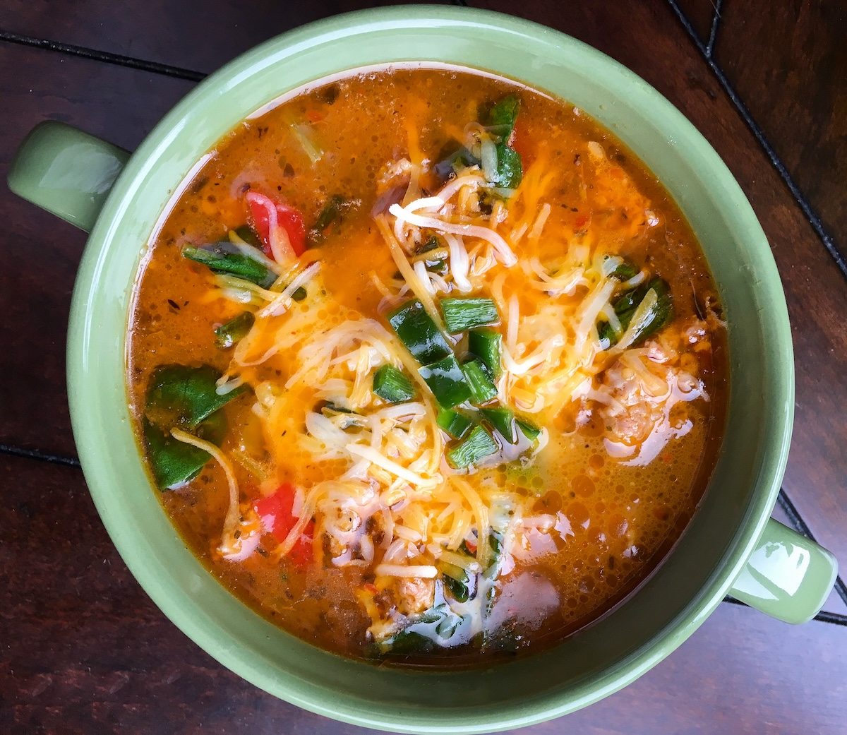 A bowl of soup with melted cheese, green beans, red bell peppers, and pieces of meat, served in a green bowl on a wooden surface.