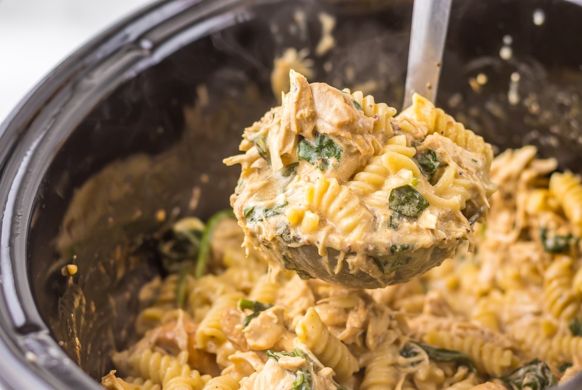 A ladle scooping creamy chicken pasta with rotini and spinach from a slow cooker.