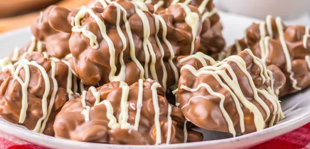 A pile of chocolate-covered nut clusters drizzled with white chocolate is arranged on a white plate.