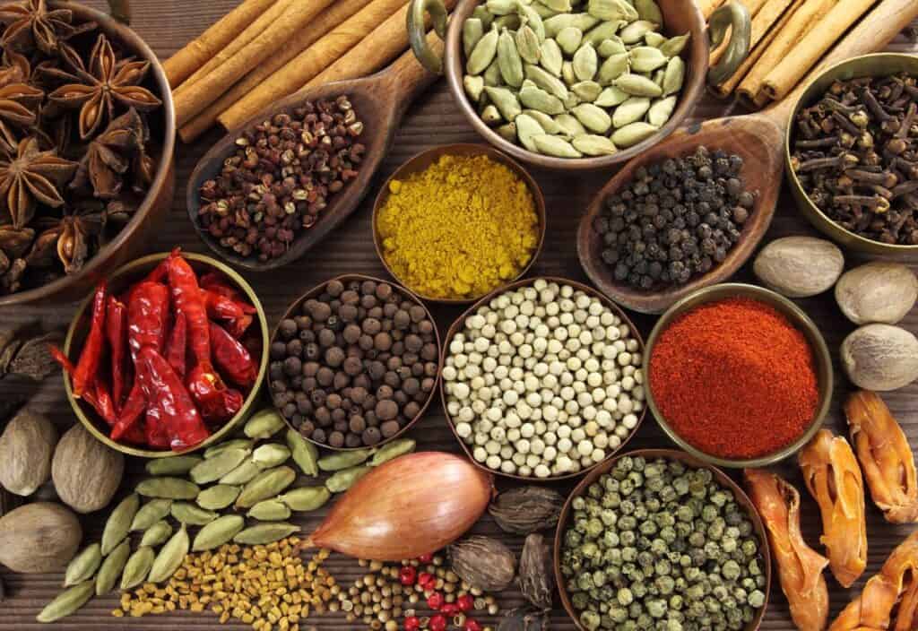 An assortment of spices and herbs in small bowls, including cinnamon sticks, star anise, cardamom pods, peppers, cloves, and chili peppers, arranged on a wooden surface.