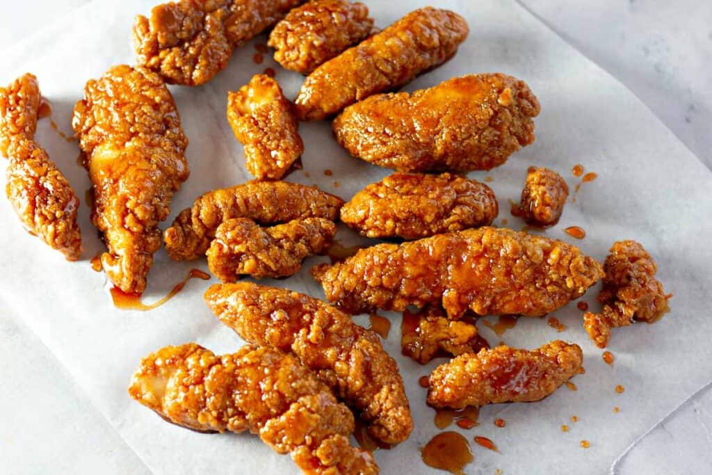 A dozen crispy, golden-brown chicken tenders on white parchment paper, with a glaze drizzled over them.