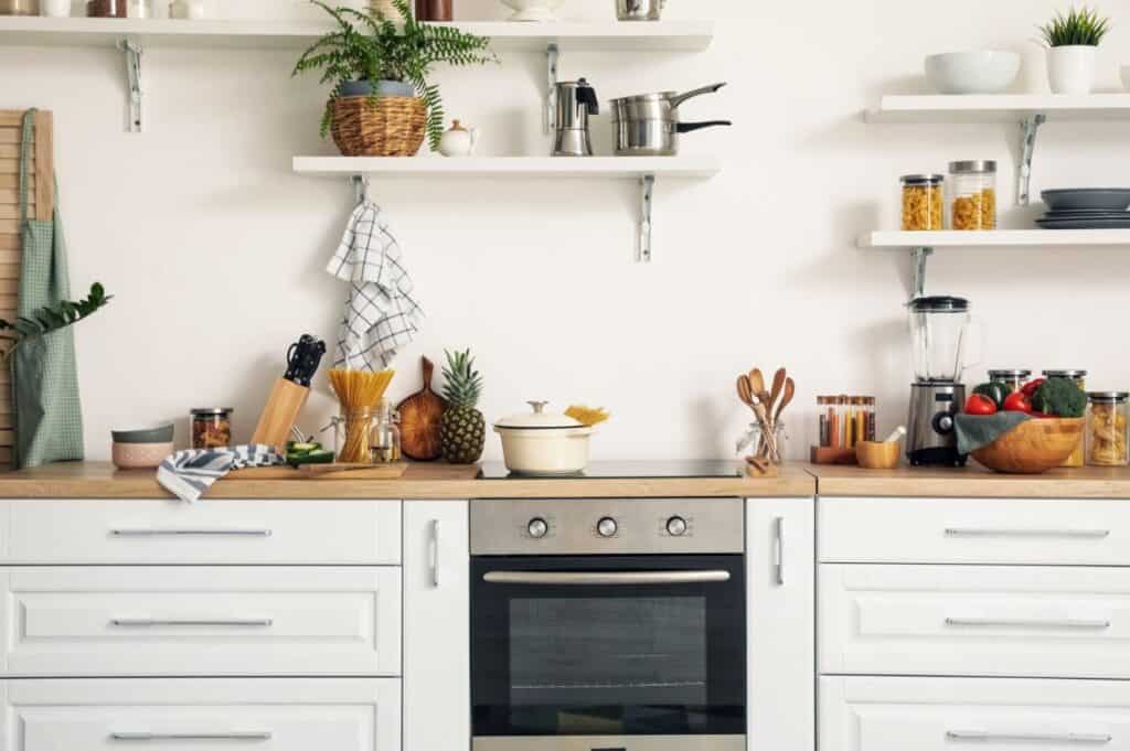 Modern kitchen refresh with white cabinets, wooden countertop, oven, and open shelves. Items include cookware, utensils, a blender, plants, and various foods like pasta, fruits, and vegetables.