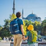 Two people with backpacks stand facing a large mosque with minarets, surrounded by palm trees and greenery on a sunny day.