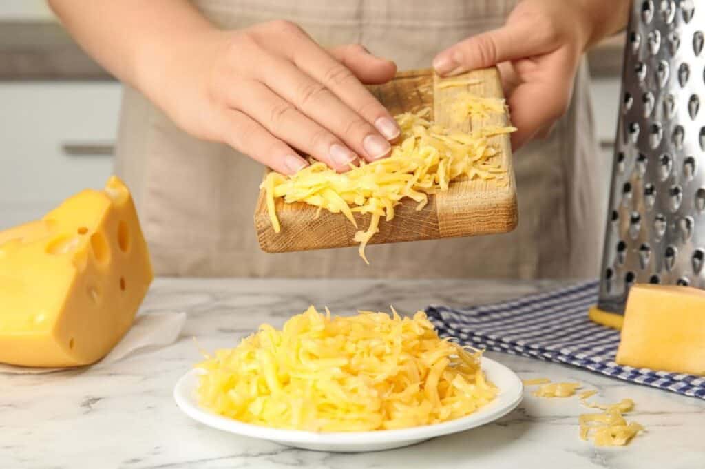 Person grating cheese over a wooden board, with shredded cheese piled on a plate. There's a cheese block and grater nearby, set on a marble countertop.