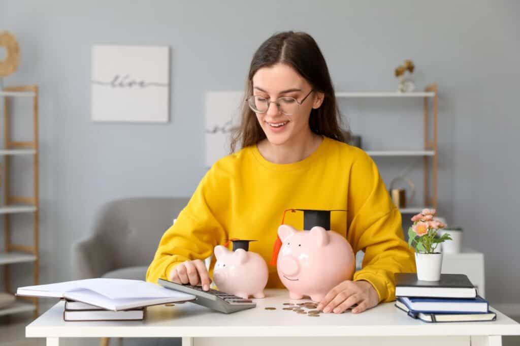 A woman in a yellow sweater counts coins at a desk with two piggy banks, one wearing a graduation cap, and some books.