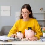 A woman in a yellow sweater counts coins at a desk with two piggy banks, one wearing a graduation cap, and some books.