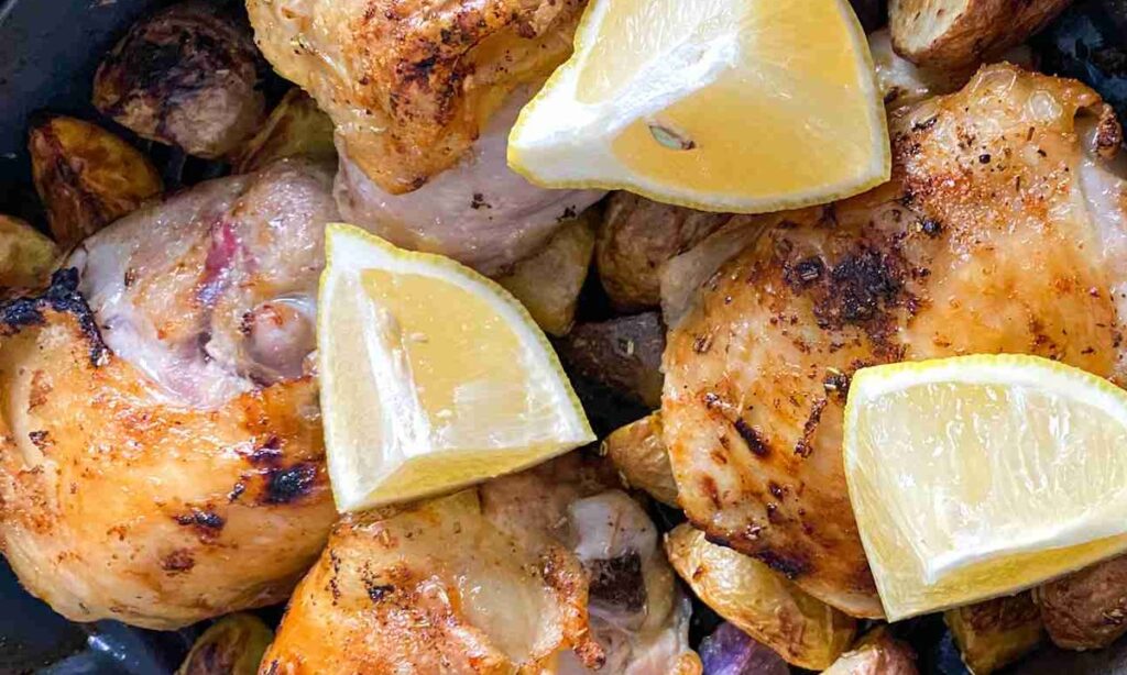 Roasted chicken thighs with crispy skin, garnished with lemon wedges, served on a bed of roasted potatoes.