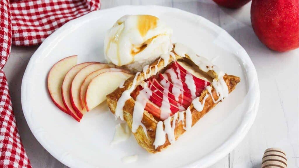 A plated dessert with a pastry topped with sliced apples and white drizzle, accompanied by a scoop of vanilla ice cream and apple slices.