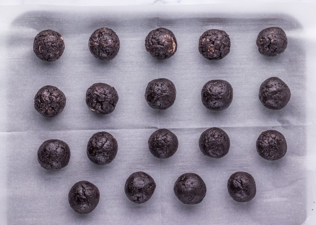 Eighteen round chocolate cookie dough balls are neatly arranged in rows on a sheet lined with parchment paper, ready for baking.