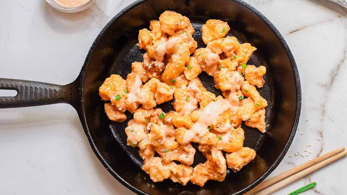 A black skillet contains pieces of breaded cauliflower drizzled with a creamy sauce, garnished with green herbs.