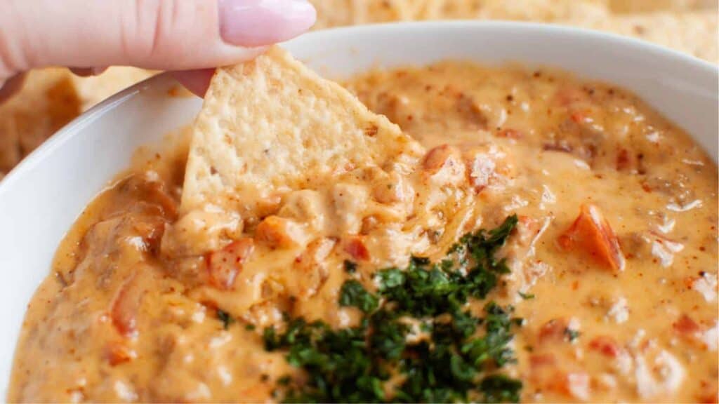 A hand is dipping a tortilla chip into a bowl of creamy cheese dip topped with herbs and visible tomato pieces.