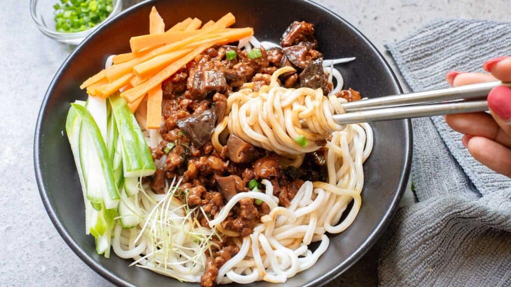 A bowl of noodles with sliced carrots, cucumbers, sprouts, and seasoned meat. A hand is using chopsticks to lift some noodles. A small dish of chopped green onions is in the background.