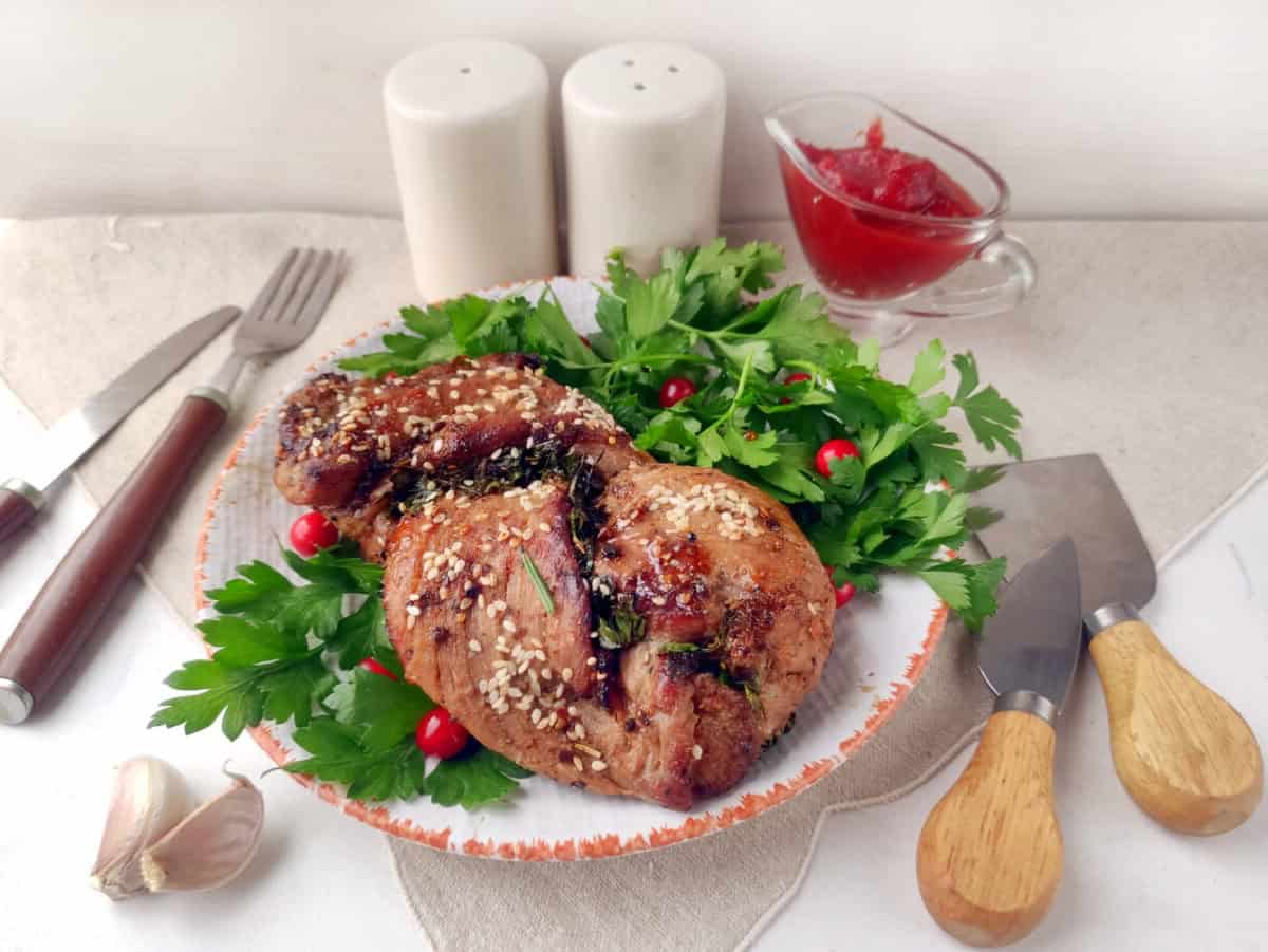 Grilled pork tenderloin with sesame seeds on a plate garnished with parsley and red berries, with garlic, cutlery, salt and pepper shakers, and a small gravy boat of sauce nearby.