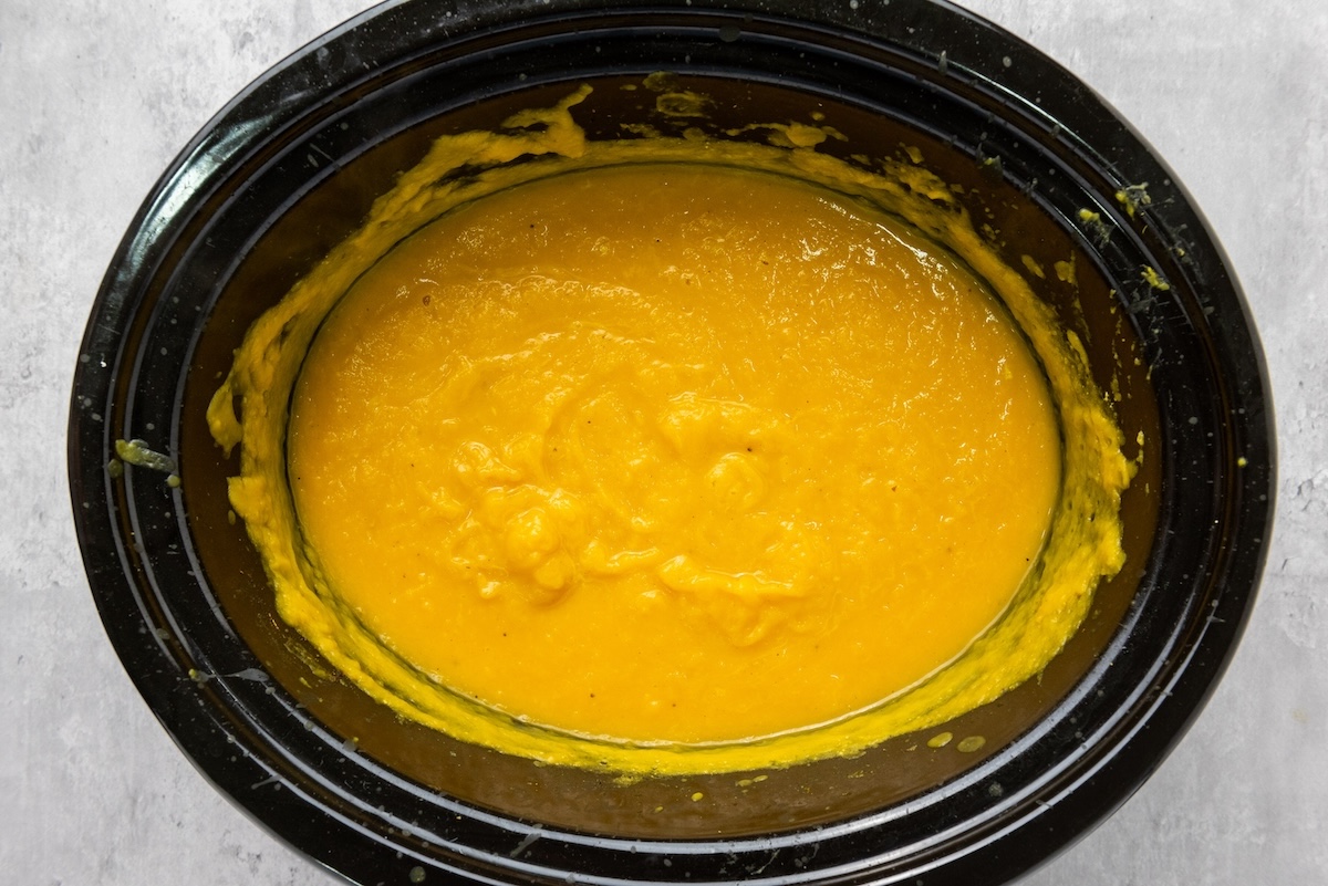 A black slow cooker filled with smooth, orange butternut squash soup on a light gray surface.