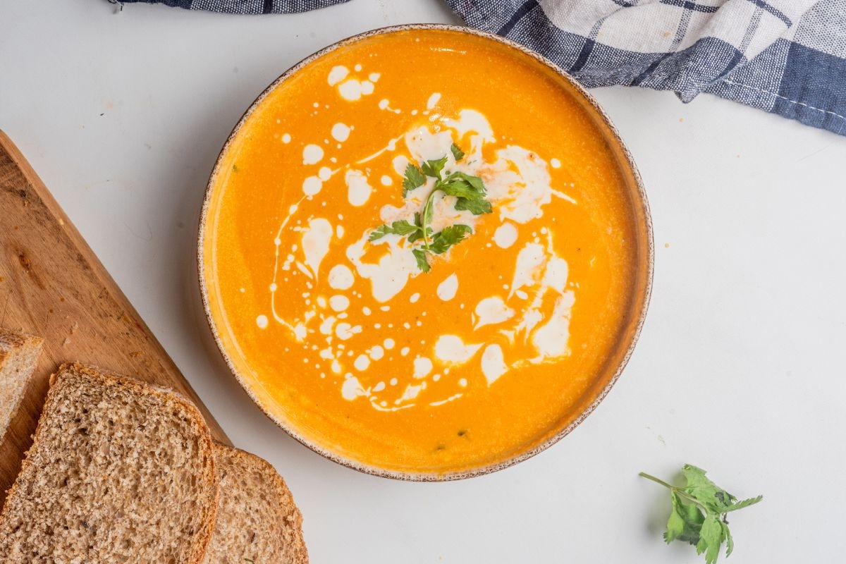 A bowl of orange soup garnished with cream and herbs is on a table next to slices of brown bread and a blue checkered cloth.