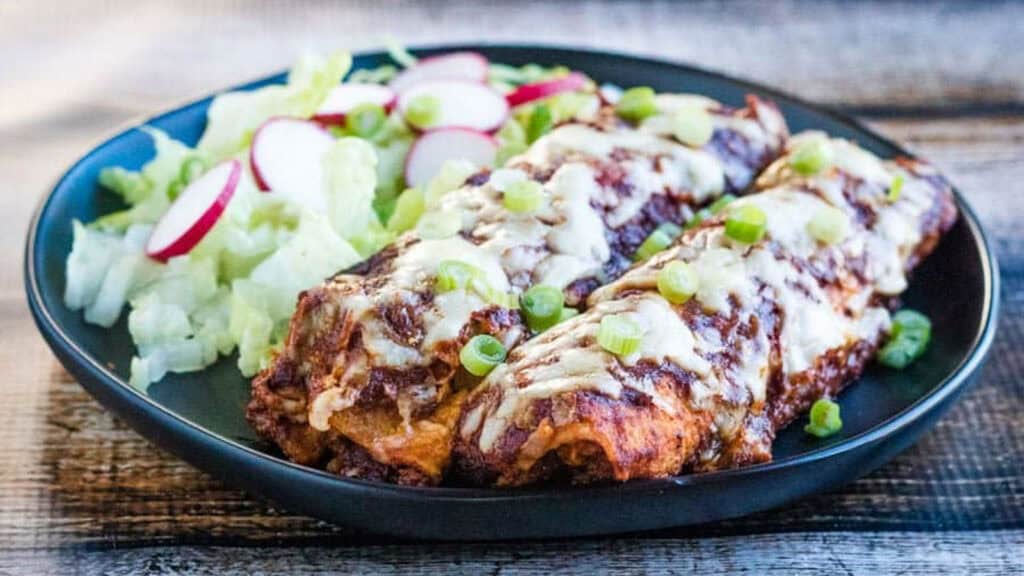 A black plate with two enchiladas topped with melted cheese and green onions, served alongside a salad with lettuce and radish slices.