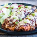 A black plate with two enchiladas topped with melted cheese and green onions, served alongside a salad with lettuce and radish slices.