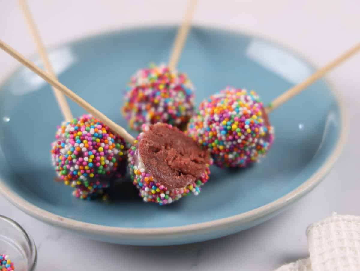 Four cake pops covered in colorful sprinkles on a blue plate, with one cake pop partially eaten to reveal the inside. Skewers are used as handles.