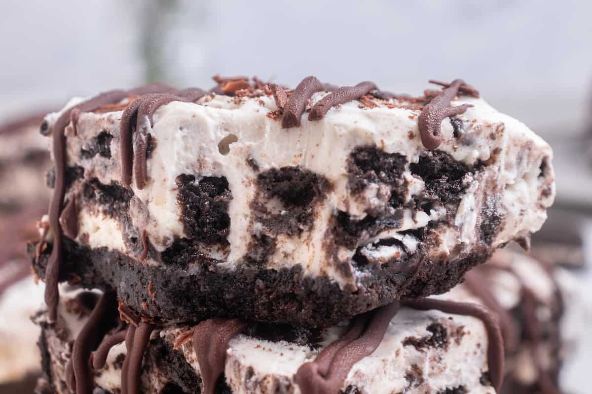 Close-up of a dessert bar with layers of cream and chocolate cookie pieces, topped with chocolate drizzle.