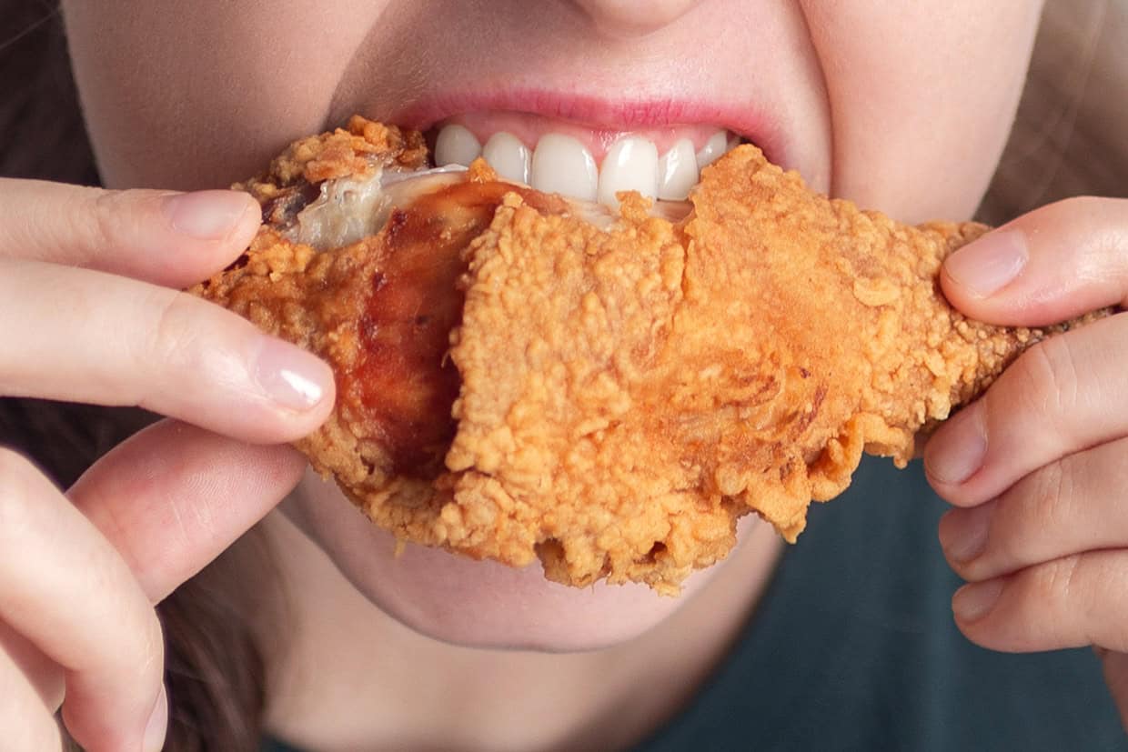 A person bites into a piece of crispy fried chicken, held with both hands, showing teeth on the chicken.