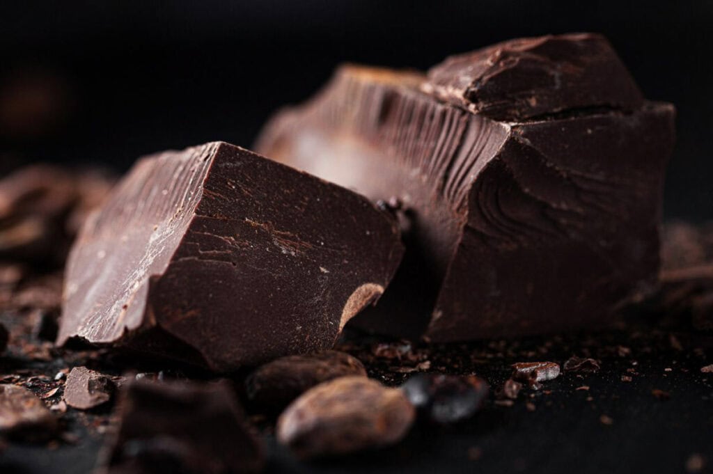 Close-up of two chunks of dark chocolate on a dark surface, surrounded by scattered cocoa nibs.