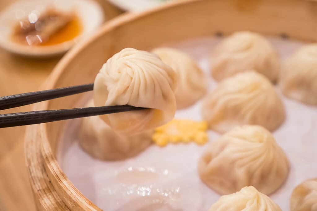 Chopsticks holding a steamed dumpling above a bamboo steamer filled with more dumplings. A small dish with sauce is in the background.