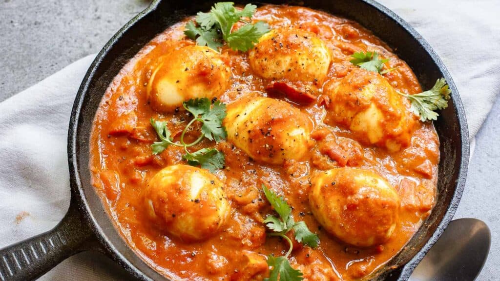 A skillet filled with spicy egg curry garnished with cilantro, featuring boiled eggs in a rich, red tomato-based sauce.