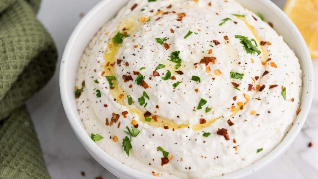 A bowl of creamy dip topped with olive oil, chopped herbs, and red pepper flakes. A green cloth and a lemon wedge are partially visible in the background.