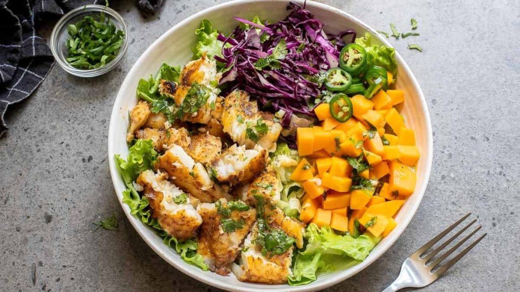 A bowl with grilled fish, chopped mango, shredded red cabbage, jalape&ntilde;o slices, and lettuce, garnished with herbs. A fork and a small bowl of chopped herbs are nearby.