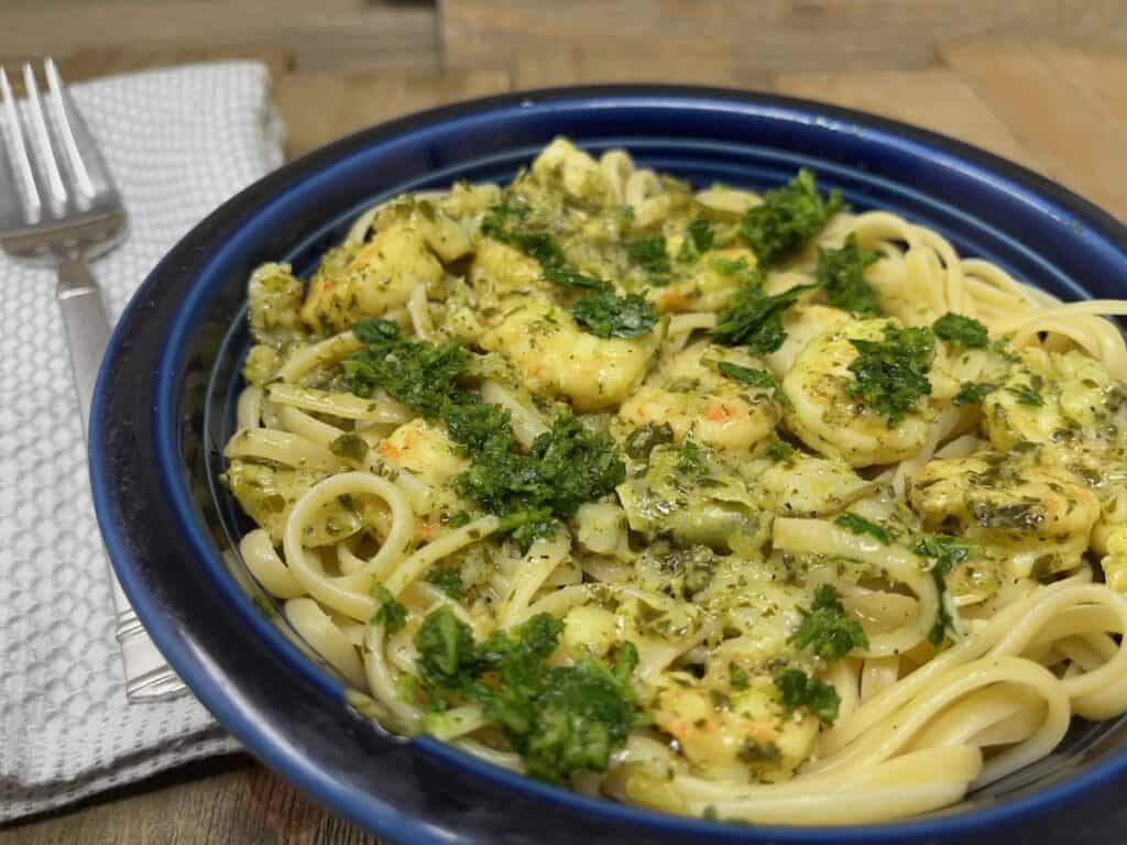 A plate of linguine with shrimp and pesto sauce, garnished with parsley. A fork and napkin are placed beside the dish.