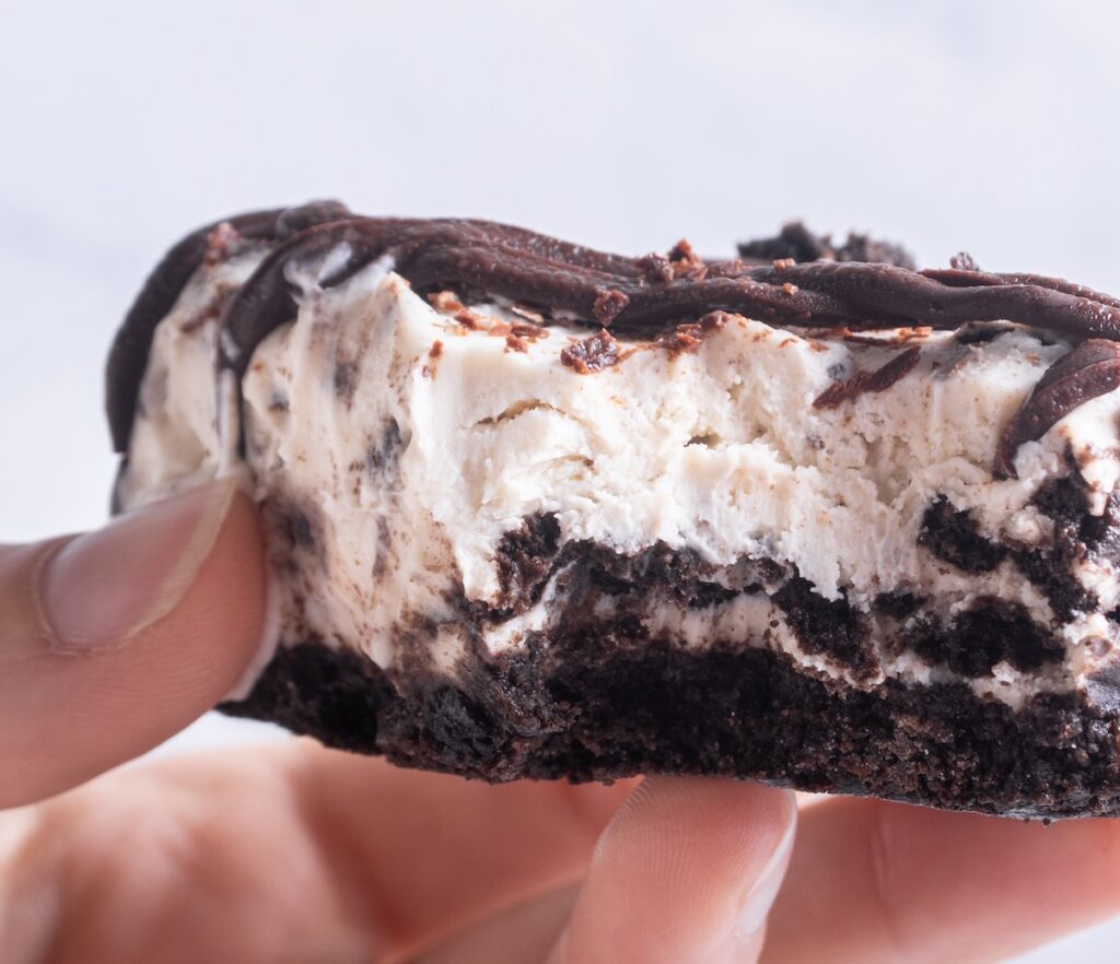 A person holds a dessert with a chocolate base, creamy filling, and chocolate drizzle. A bite has been taken from it.