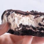 A person holds a dessert with a chocolate base, creamy filling, and chocolate drizzle. A bite has been taken from it.