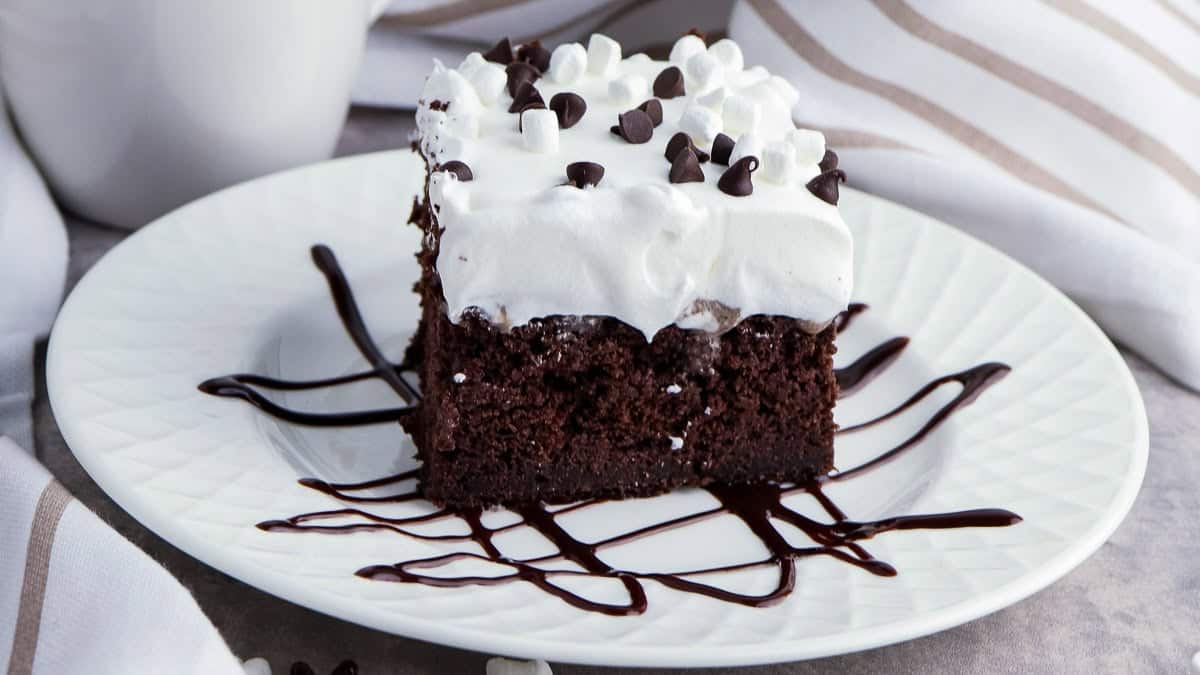 A slice of chocolate cake with white frosting and chocolate chips on top, drizzled with chocolate syrup on a white plate.