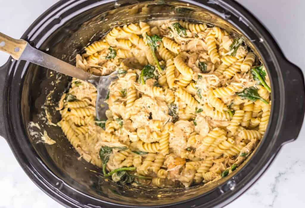 A crockpot filled with cooked rotini pasta mixed with spinach, shredded chicken, and a creamy sauce, with a serving spoon inside.