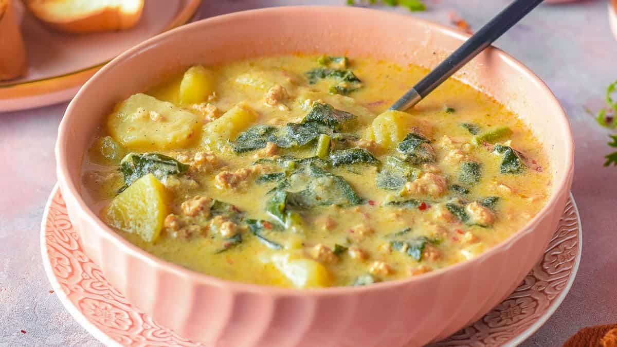 A bowl of creamy yellow soup with chunks of sausage, potato and leafy greens.