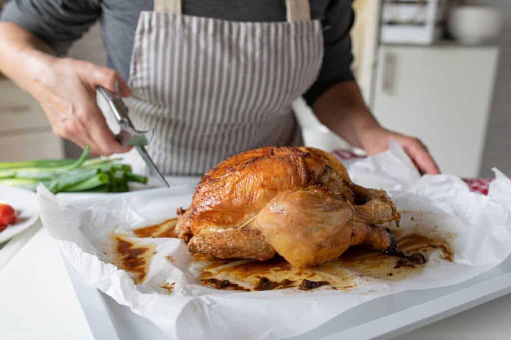 Person carving a roasted chicken on a tray in a kitchen, with green onions nearby.