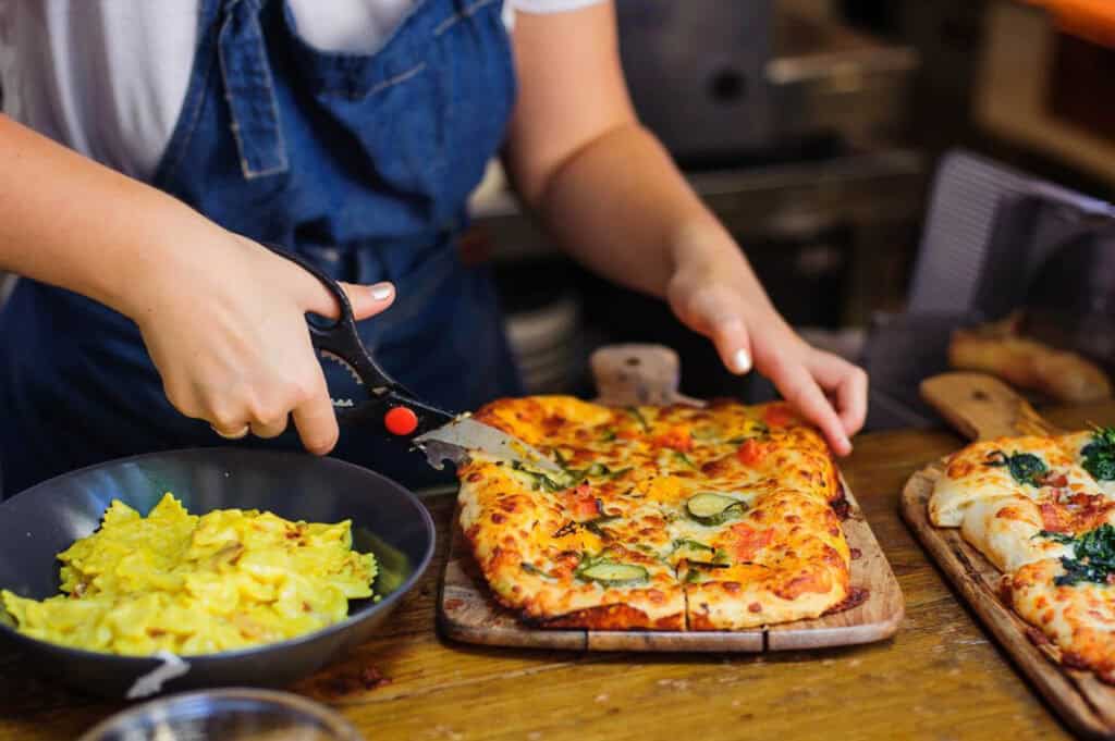 A person in a blue apron cuts a rectangular pizza with scissors. A bowl of pasta is nearby on a wooden table.