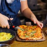 A person in a blue apron cuts a rectangular pizza with scissors. A bowl of pasta is nearby on a wooden table.