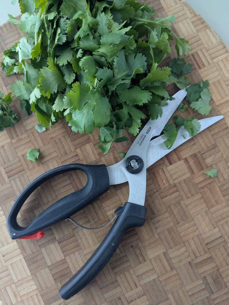 Kitchen scissors on a wooden cutting board, next to a pile of fresh cilantro leaves.