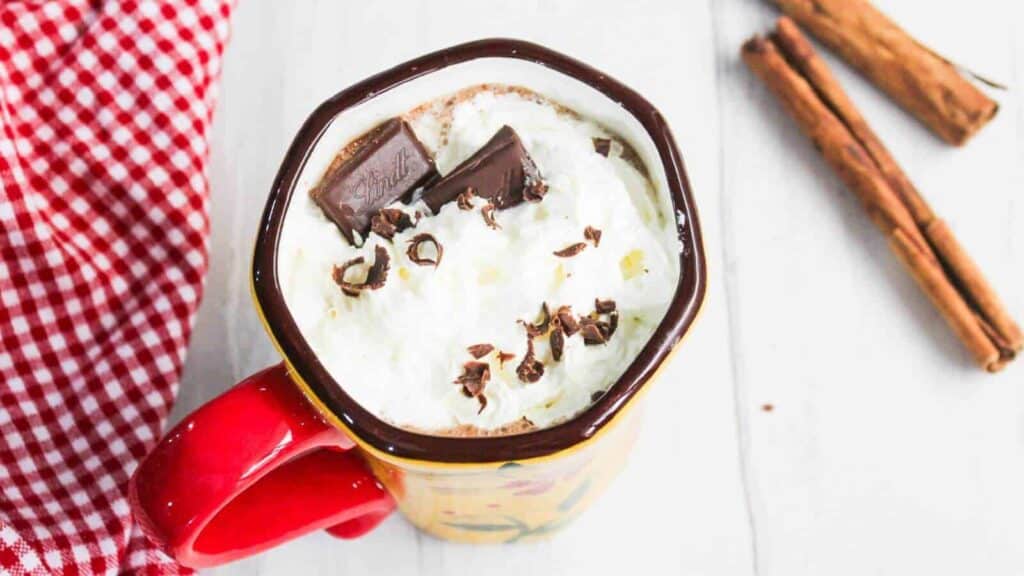 A mug of hot chocolate topped with whipped cream, chocolate shavings, and a chocolate piece. A red checkered cloth and cinnamon sticks are nearby.