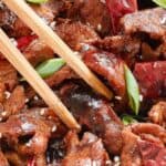 Close-up of a dish featuring stir-fried beef with dried red chili peppers, garnished with green onions and sesame seeds. Chopsticks rest on top.