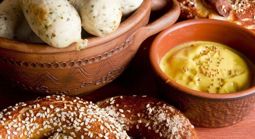 A ceramic bowl of sausages, a pretzel topped with sesame seeds, and a small bowl of mustard with seeds on top.