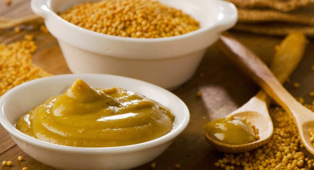 A bowl of mustard paste next to a bowl of mustard seeds with a wooden spoon on a wooden surface.