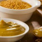 A bowl of mustard paste next to a bowl of mustard seeds with a wooden spoon on a wooden surface.